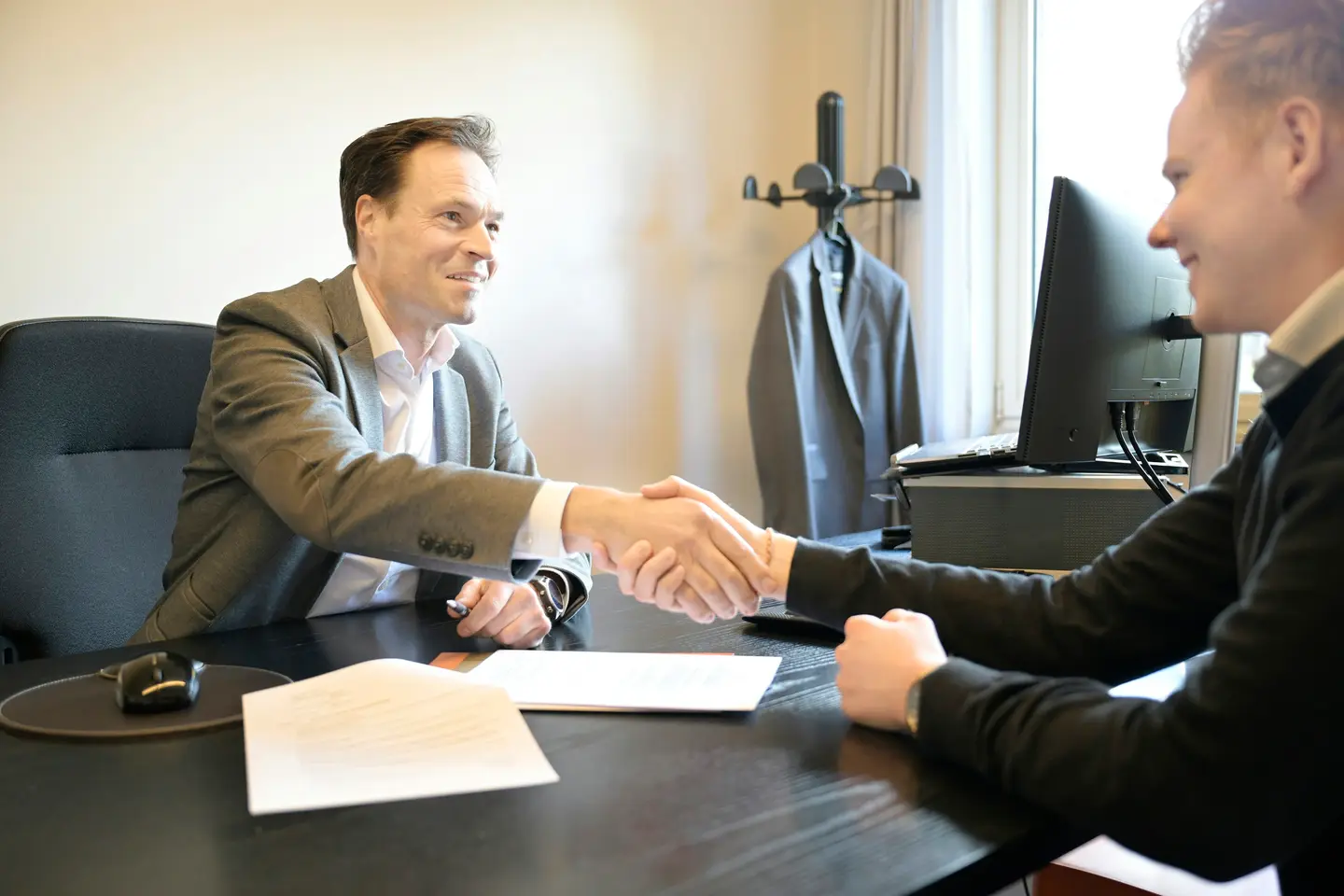 Zwei Männer in Hemd und Sakko geben sich über einem Schreibtisch lächelnd die Hand